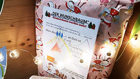 Das nolteteam ist wieder dabei bei der Aktion Emsdettener Wunschbaum Rosa eingepacktes Geschenk mit einem Zettel drauf, der eine Kinderzeichnung einer Prinzessin zeigt und einen Text zum Wunschbaum. Davor eine Lichterkette.