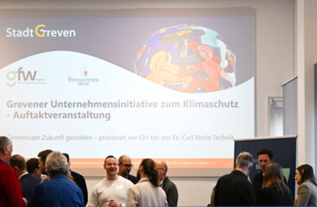 Auf einer Beamerleinwand ist ein Chart der Veranstaltung "Grevener Unternehmensinitiative zum Klimaschutz - Auftaktveranstaltung" dargestellt. Davor zu sehen einige Personen, die miteinander im Gespräch sind.